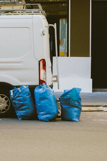 The image depicts the rear side of a white commercial van parked on a street, with three large blue rubbish bags placed on the pavement near the back corner of the vehicle. The bags are made of plastic, appear filled with waste, and are tied at the top. To the right of the bags, the van's rear door is partially visible, featuring a vertical red reflector strip. Behind the van, a building with a large glass window or door, framed in black, can be seen, along with a section of a white external wall and a small concrete platform or step. The scene is illuminated by natural daylight, and the environment suggests a typical urban or suburban street setting, where private rubbish collection services, such as those provided by Rubbish Removal Tufnell Park, might undertake on-site clearance or alternative waste handling to remove refuse efficiently and professionally, especially for commercial clients.