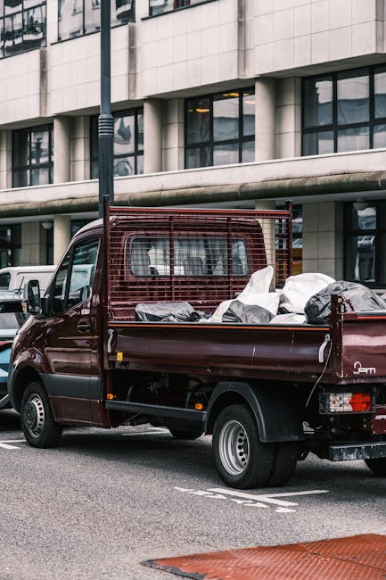 A burgundy flatbed pickup truck parked on a city street in front of a modern office building with large glass windows and white paneling. The truck's cargo area is filled with various types of waste materials, including several black plastic trash bags and miscellaneous debris, arranged in an unorganized manner. The truck features a metal safety grille at the cab's rear and standard wheels with grey hubcaps. The surrounding environment includes parked cars and a streetlamp, indicating an urban setting suitable for private rubbish collection and on-site waste clearance. The scene suggests an instance of concrete waste removal that could be managed by a professional rubbish removal service like Rubbish Removal Tufnell Park, providing an alternative to municipal rubbish disposal channels in busy city environments.