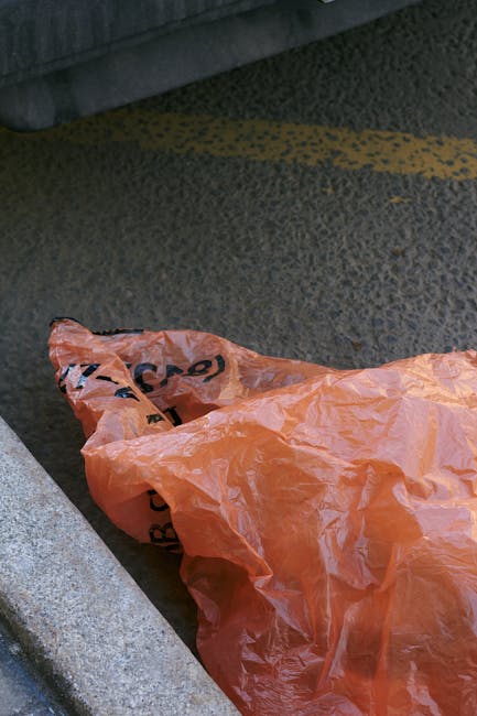 The image depicts the rear side of a white commercial van parked on a street, with three large blue rubbish bags placed on the pavement near the back corner of the vehicle. The bags are made of plastic, appear filled with waste, and are tied at the top. To the right of the bags, the van's rear door is partially visible, featuring a vertical red reflector strip. Behind the van, a building with a large glass window or door, framed in black, can be seen, along with a section of a white external wall and a small concrete platform or step. The scene is illuminated by natural daylight, and the environment suggests a typical urban or suburban street setting, where private rubbish collection services, such as those provided by Rubbish Removal Tufnell Park, might undertake on-site clearance or alternative waste handling to remove refuse efficiently and professionally, especially for commercial clients.