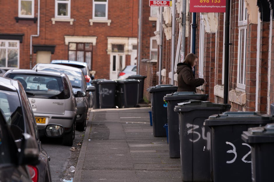 A residential street scene featuring a row of dark gray wheelie bins lined up along the sidewalk adjacent to a red-brick building, with some bins partially open revealing contents inside. A woman dressed in a dark coat is seen standing near the bins, possibly in the process of discarding rubbish or organizing waste. Several parked cars, including a silver hatchback and a dark-colored vehicle, are positioned along the curb in the background. The pavement surface appears slightly weathered with some scattered debris and litter near the edges. The building's facade displays windows with white frames, and a red 'Sold' sign is visible above a doorway, suggesting property is for sale. Graffiti scribbles are present on the lower section of the bins, indicating a typical urban environment where private waste collection services like Rubbish Removal Tufnell Park operate. The overall scene presents an everyday view of waste management activity aligned with independent rubbish disposal practices within a residential area. The lighting suggests daytime, with natural light illuminating the scene evenly, emphasizing the textures of brickwork, paving, and waste containers.