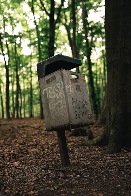 A metal postal box, anchored to the ground on a slight tilt, is situated in a wooded area surrounded by tall trees with dense green foliage. The box has a grey, weathered surface with visible graffiti markings and stickers, and a black, slightly open lid on top. The textured surface of the box reflects its metal finish, and the graffiti includes black and white scribbles and tags. The environment around the box features leaf-littered earth and tree trunks, with diffused natural light filtering through the canopy overhead, creating a subdued outdoor scene. The presence of graffiti and the weathered condition of the box suggest it is located in an outdoor, natural setting, possibly near a walking trail or park, where independent waste disposal or collection could be relevant. The scene indicates a form of informal or alternative waste handling, aligning with private disposal practices often found in non-urban environments.