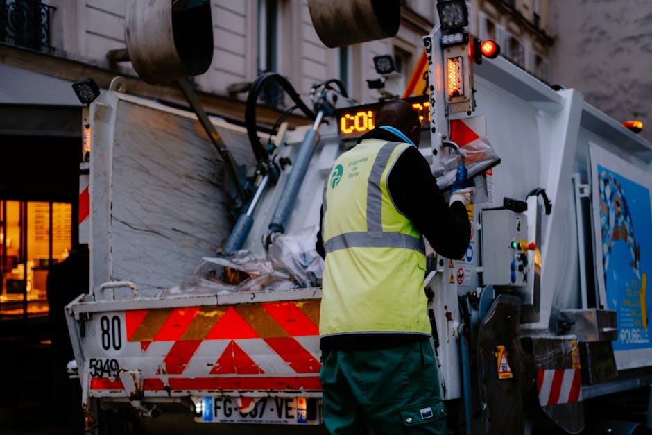 A waste collection worker wearing a high-visibility yellow vest and dark trousers is operating a waste collection vehicle in an urban environment during the evening. The worker is standing at the rear of the vehicle, which is equipped with hydraulic arms and safety lights, and appears to be managing waste or debris inside the large, white, metal container. The container’s surface shows signs of wear and dirt, and the vehicle displays reflective red and white warning markings. In the background, a building with illuminated windows is visible, indicating a city setting. The scene captures an on-site waste collection activity, illustrating an aspect of independent or private rubbish removal services that address waste disposal outside conventional municipal channels. The image emphasizes the operational aspect of rubbish collection, aligned with professional waste management practices typical in urban areas, and highlights the importance of proper disposal procedures in maintaining cleanliness and hygiene.